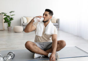 An image of a healthy man illustrated by a guy sitting cross-legged on a yoga mat, drinking water.