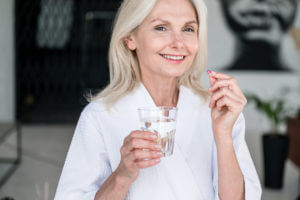 An image of a smiling mature woman holding a pill and a glass of water to portray anti-aging and human life extension.