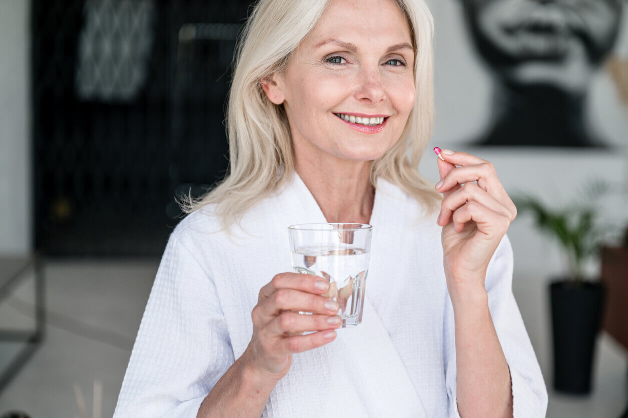 An image of a smiling mature woman holding a pill and a glass of water to portray anti-aging and human life extension.