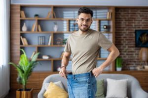 A young man is smiling at home, standing in front of the camera, wearing jeans and showing weight loss results from weekly weight loss injections.